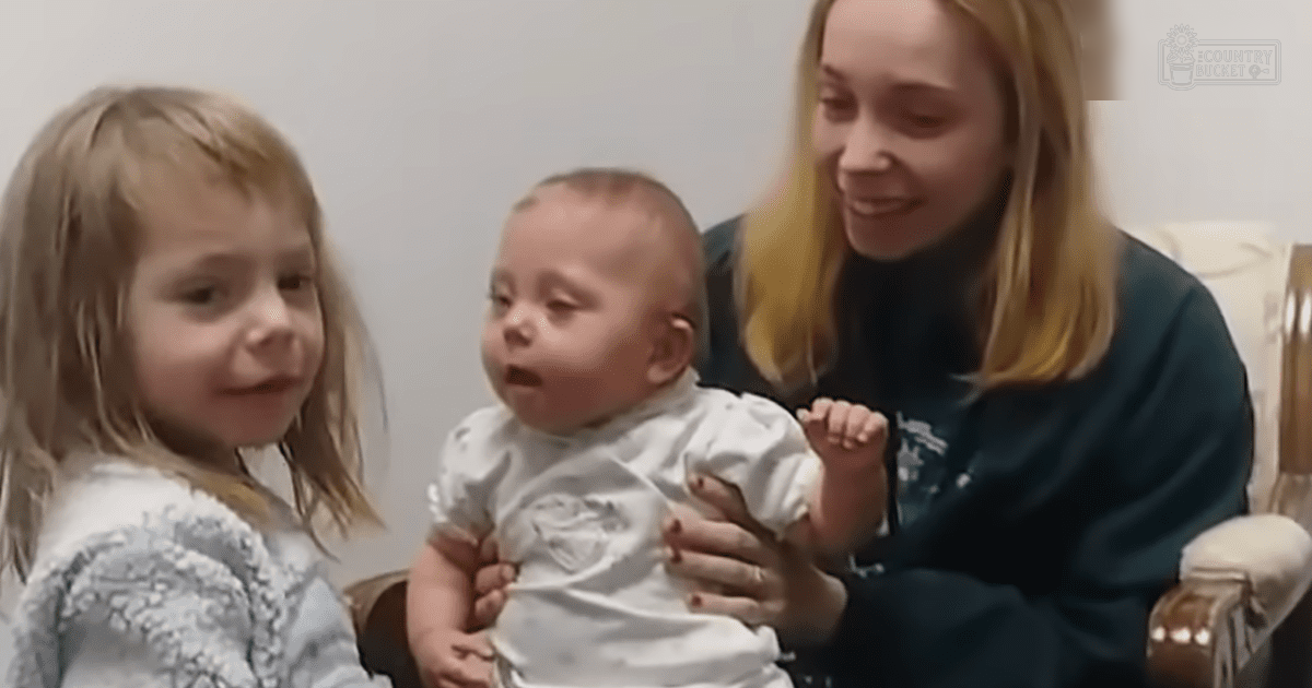A young adult female, toddler, and infant indoors with simple furniture and plain white walls.