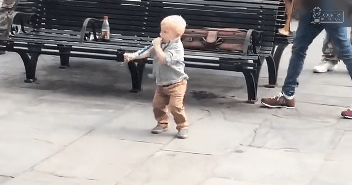 A toddler in a light gray long-sleeve shirt and brown pants stands on an urban park bench with a black metal bench and a brown suitcase behind him, surrounded by adults in jeans and sneakers; the setting is paved and appears to be daytime.