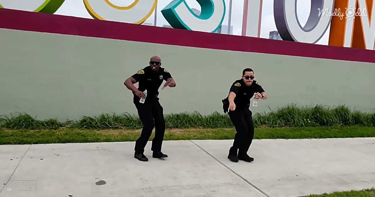 Two adults in dark uniforms dancing outdoors near a colorful sign, suggesting an event setting with partly cloudy skies.