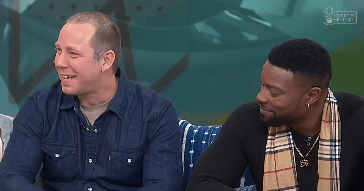 Two adult males in a studio setting, wearing casual and semi-formal attire with accessories; blue denim shirt, black ribbed sweater, plaid scarf, gold chain necklace.