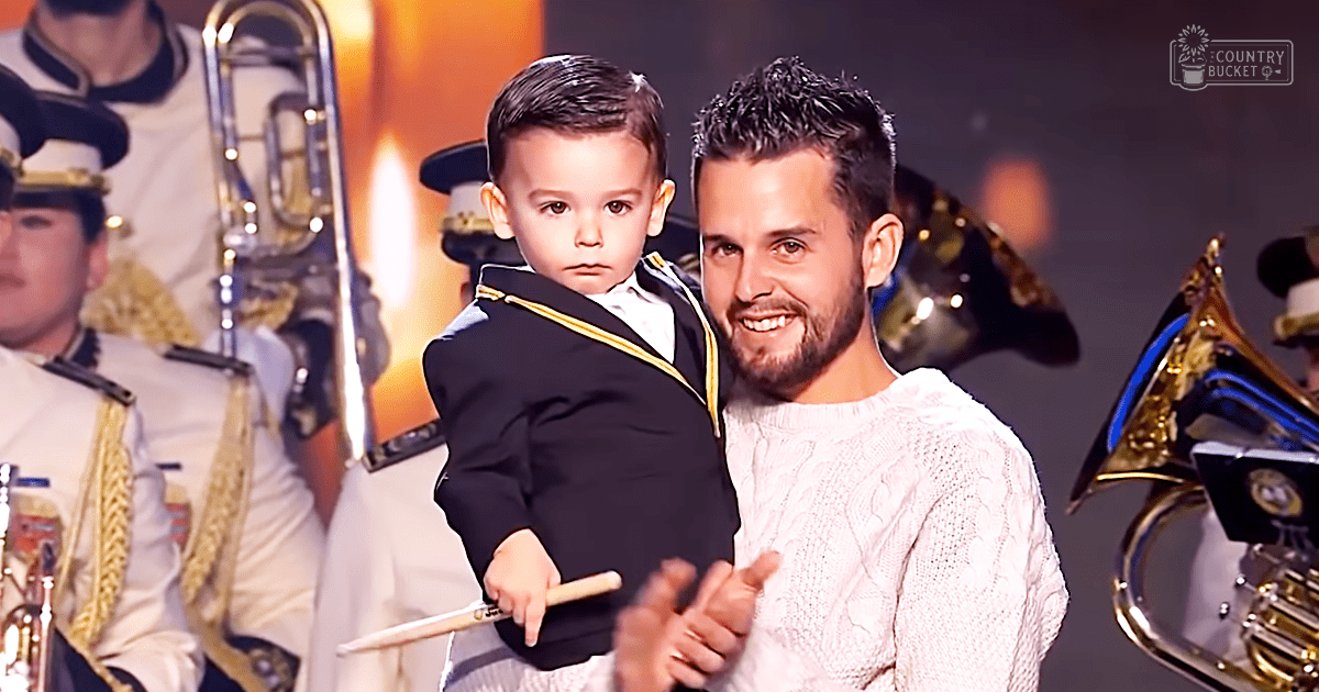A young adult male singer holds a toddler dressed formally, suggesting an indoor performance setting with brass band instruments visible. The scene is illuminated by stage lighting, typical of a concert hall or theater environment.