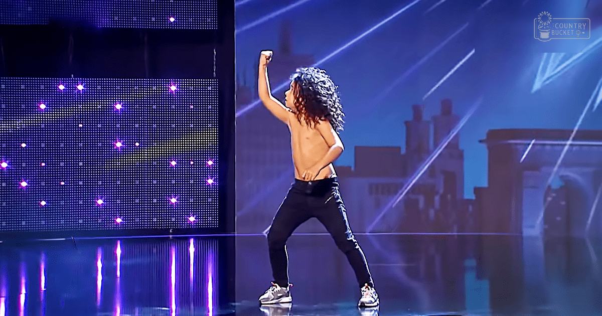 A young adult male performing on stage at a talent show with bright stage lights and an urban cityscape backdrop.