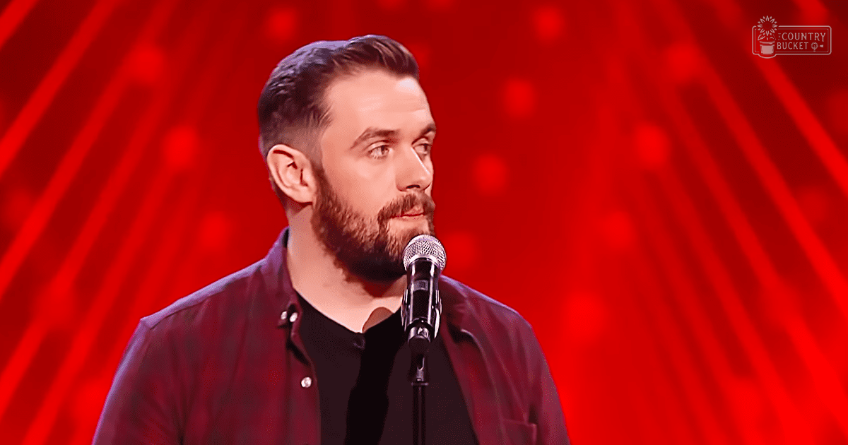 An adult male performer wearing a dark red button-up shirt and black undershirt stands on stage with professional red lighting in the background, suggesting a comedy club setting.
