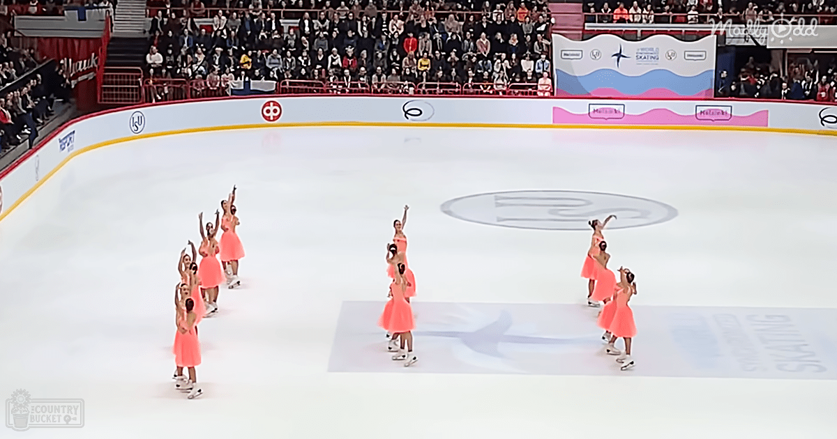 A synchronized skating team performs at an indoor ice rink, dressed in coral-colored dresses with white skates, surrounded by spectators seated in stands under well-lit artificial lighting.