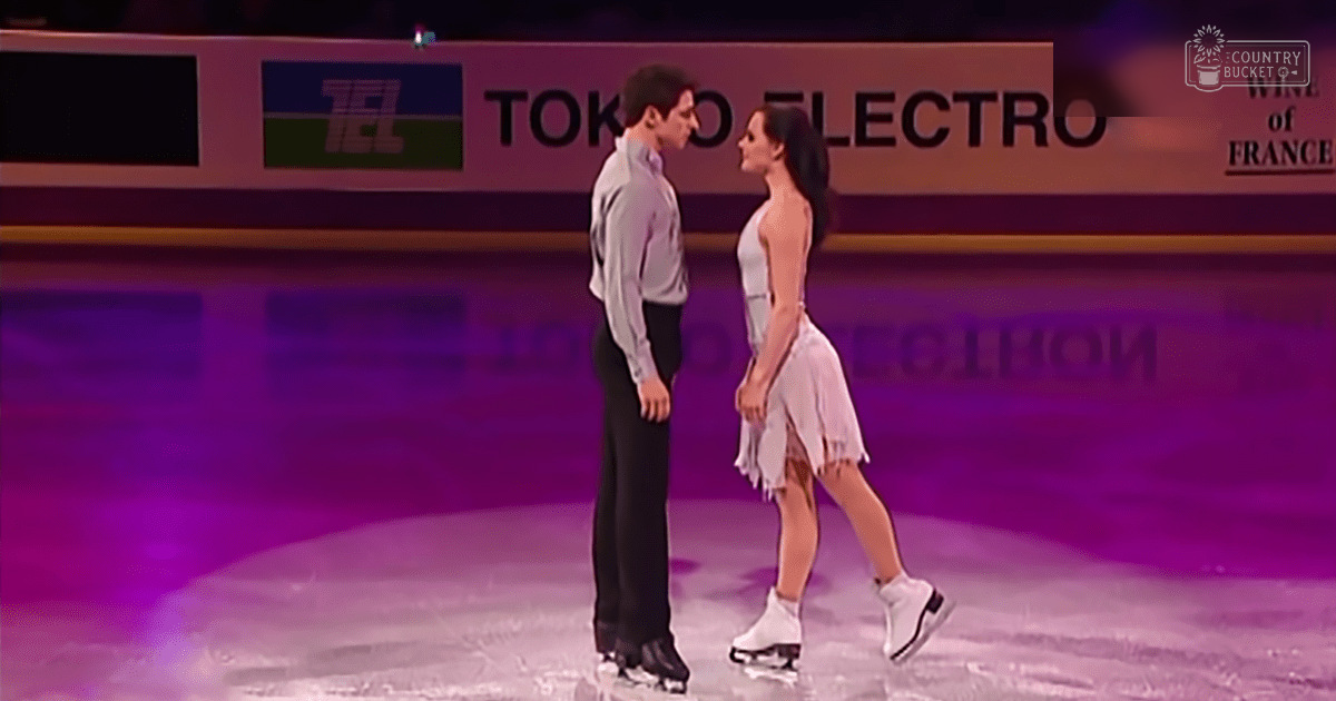 A male and female figure skating pair at an indoor ice rink, wearing light gray long-sleeved shirts and black pants for the male, white sleeveless fringed dresses with asymmetrical hemlines for the female. The setting appears professional, likely a competition or exhibition.