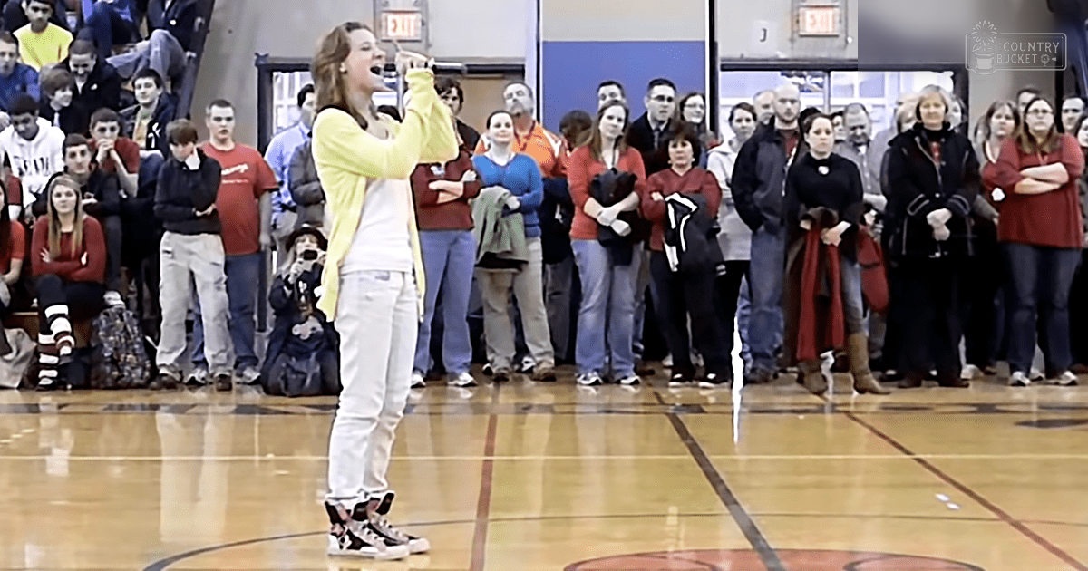 Young adult female singing in a gymnasium filled with spectators during daytime.