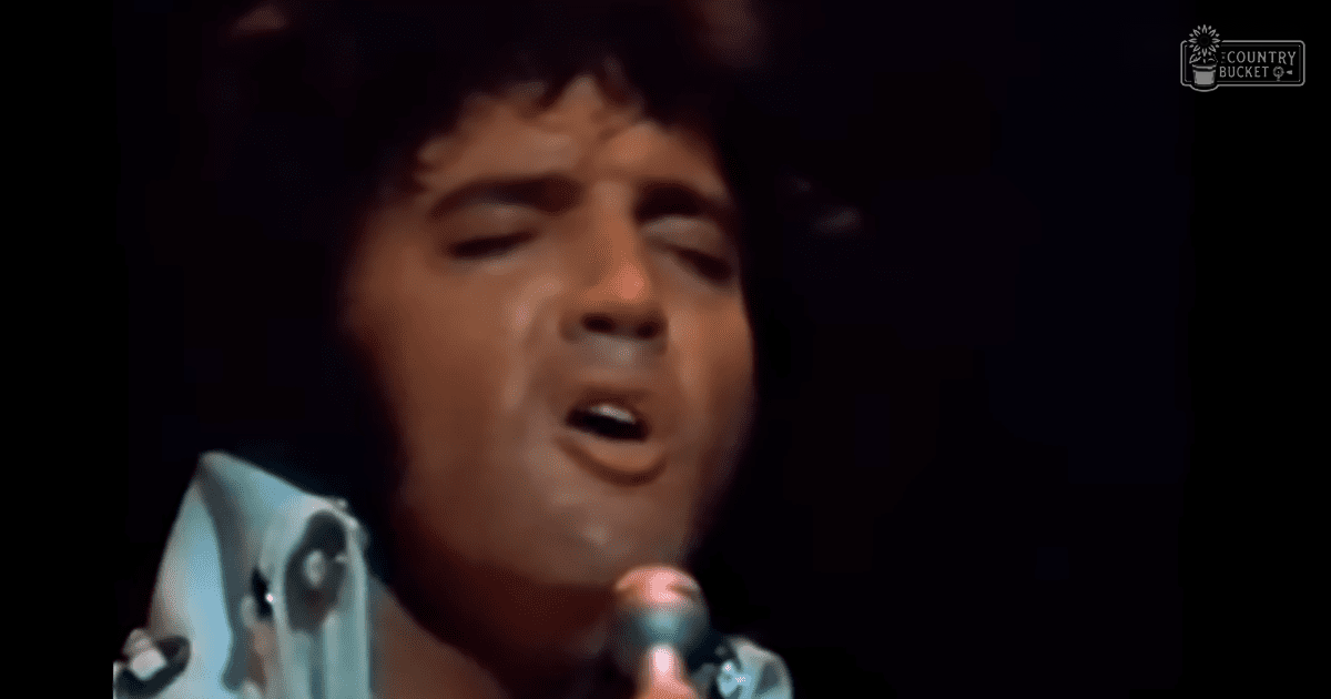 An adult male performer singing into a microphone, wearing a light blue, patterned shirt with circular designs and floral motifs in a relaxed fit, on stage under dim lighting typical of evening performances.