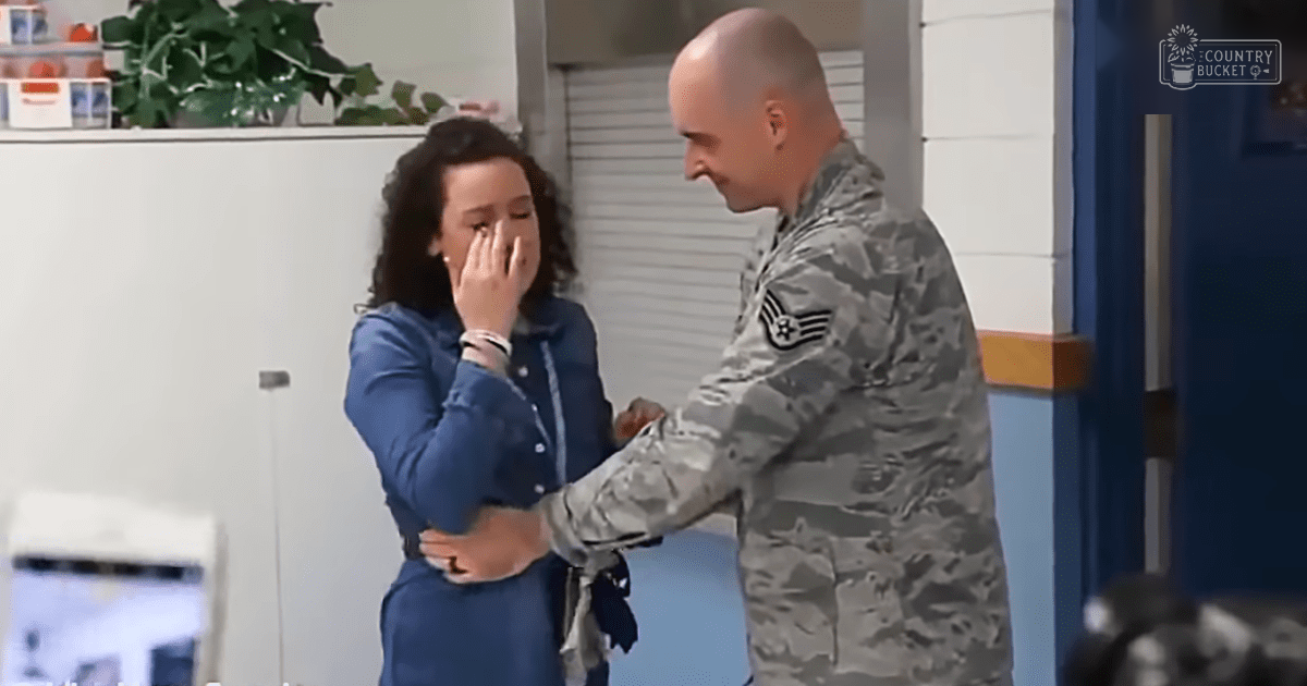 A woman in a modern professional blue button-up dress with long sleeves, tailored fit, and straight neckline stands in an indoor office environment. A man in a camouflage military uniform is also present.