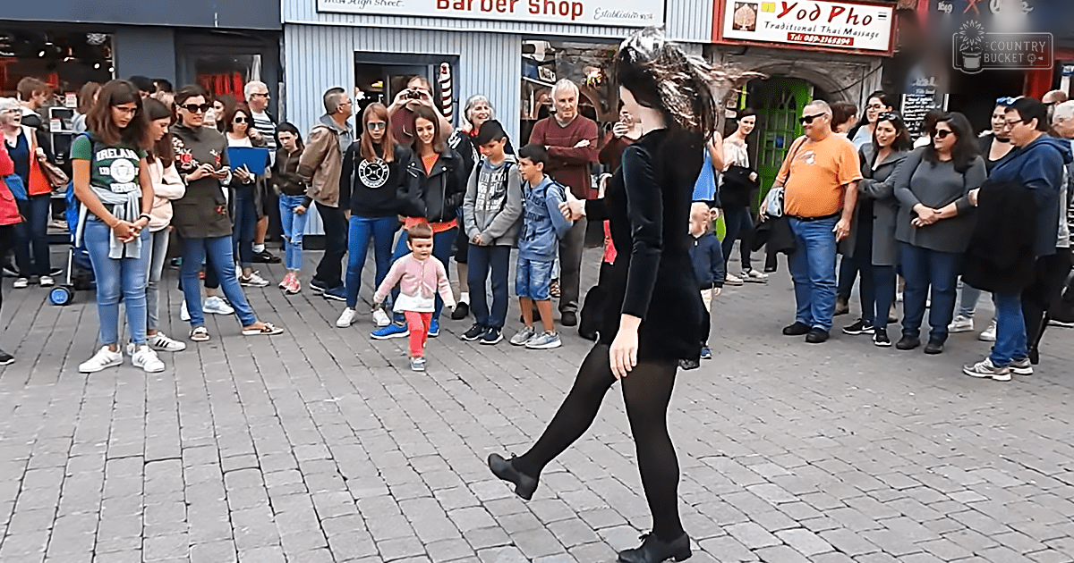 A young adult female dancer in motion on an outdoor street scene with storefronts, including a barber shop and Thai massage parlour, under natural daylight during spring or summer.