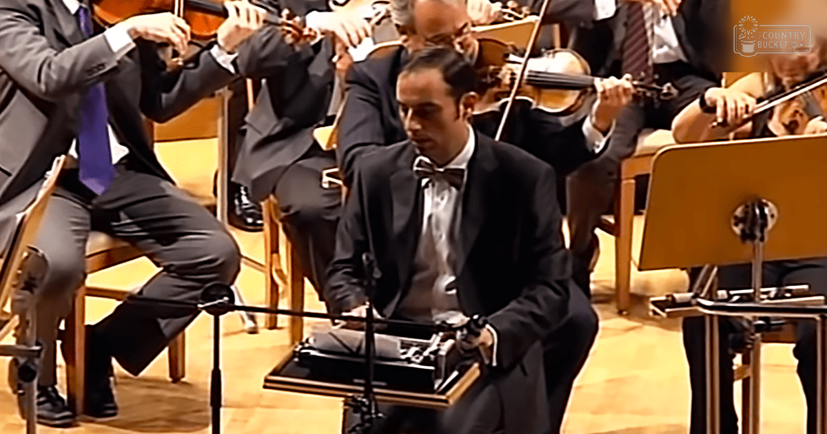 Adult male conductor in an orchestra setting, wearing a black tuxedo with a white dress shirt and a black bow tie. Other musicians are seated behind him, playing violins.