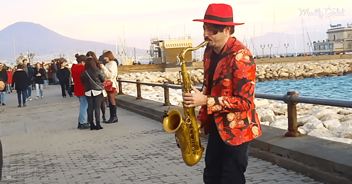 An adult male singer plays a gold saxophone outdoors near a waterfront promenade with people walking and boats docked at a pier under partly cloudy skies.