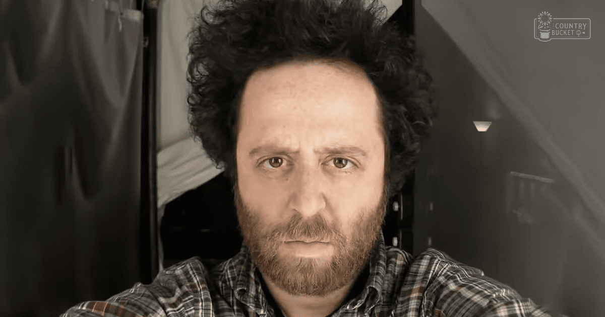 Middle-aged adult male wearing a casual plaid shirt indoors with natural light and black curtains behind him.