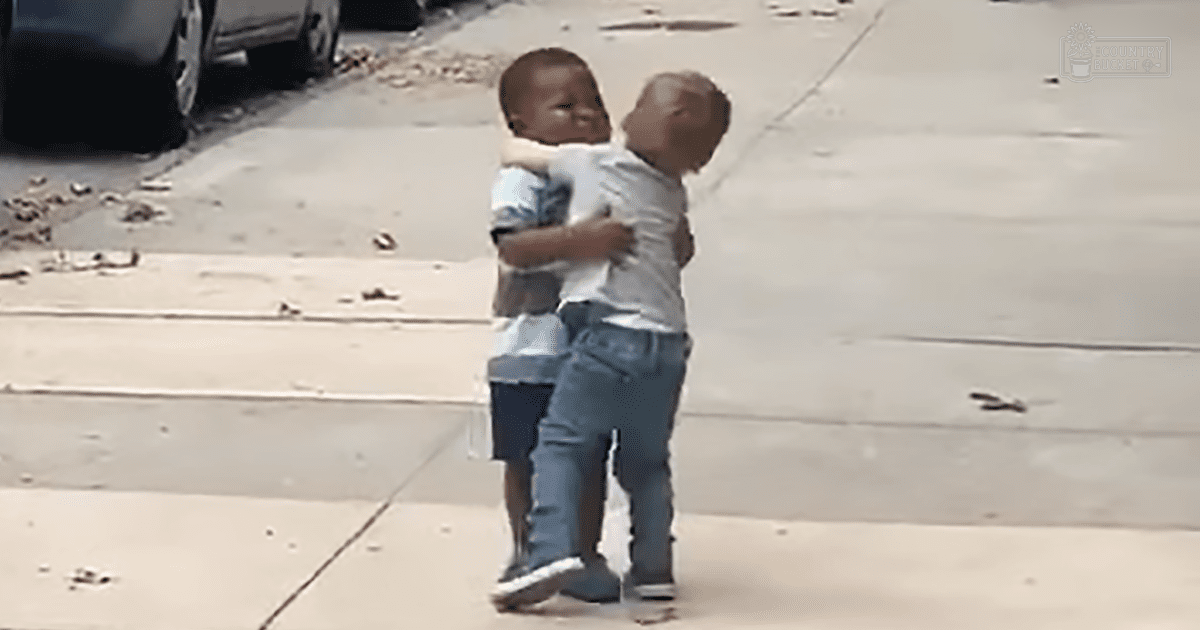 Two toddlers hugging each other outdoors on a sidewalk with scattered debris, during daytime light.