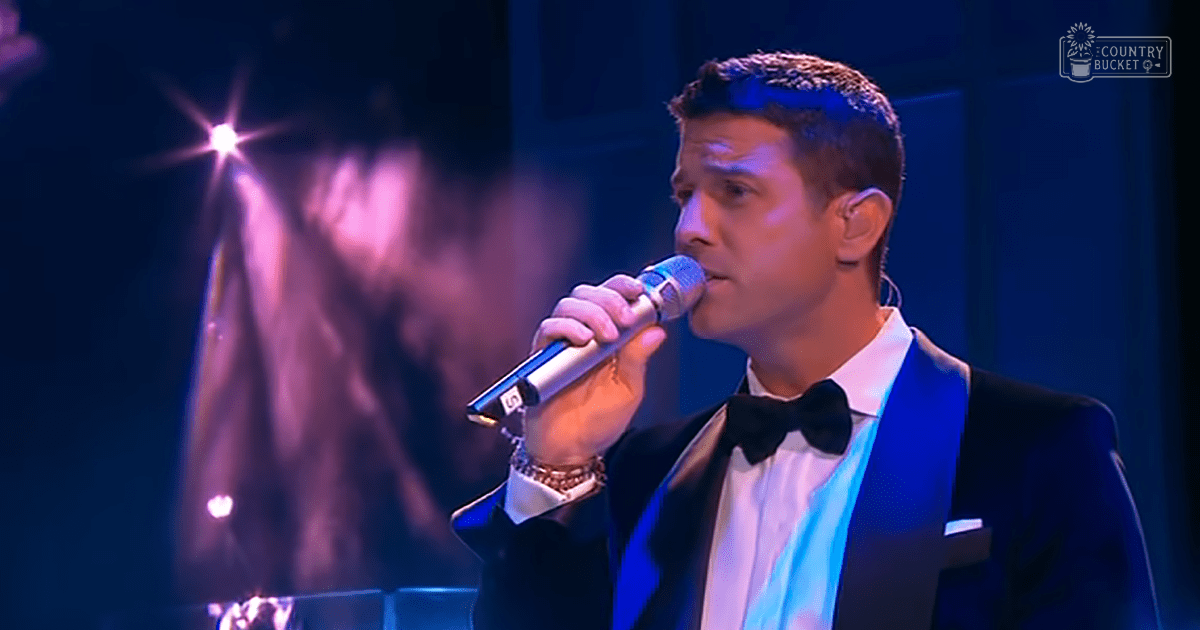 An adult male singer in a black tuxedo with white shirt and black bow tie performs into a microphone at what appears to be an indoor concert setting, likely a theater or hall.