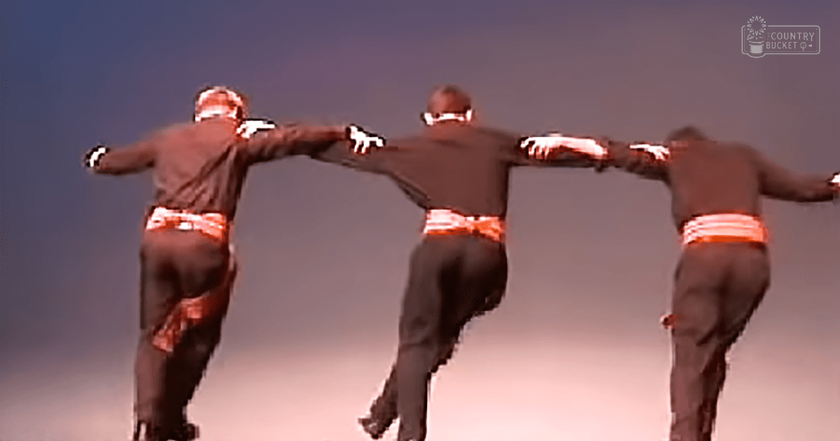 A group of male performers in tight-fitting pants with orange and white stripes, loose-fitting brown shirts, and black gloves against a gradient sky transitioning from dusk to night.