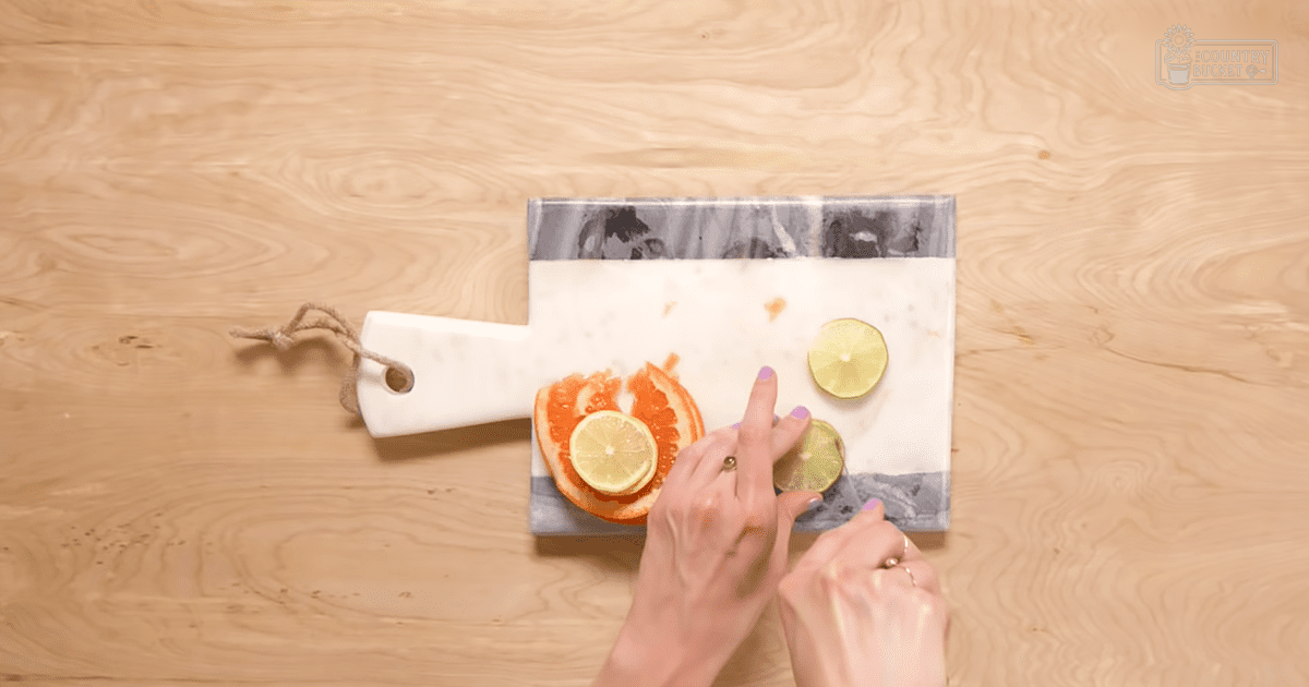 A woman, wearing rings, prepares sliced grapefruit and lemon on a white marble cutting board with grayish-blue border in an indoor kitchen setting under bright even lighting.
