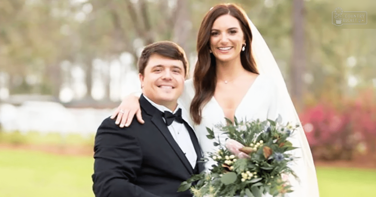 A young adult male singer in a formal black tuxedo stands beside an adult female wearing a white wedding dress with long sleeves, holding a bouquet of greenery and small white flowers at an outdoor garden or park setting.