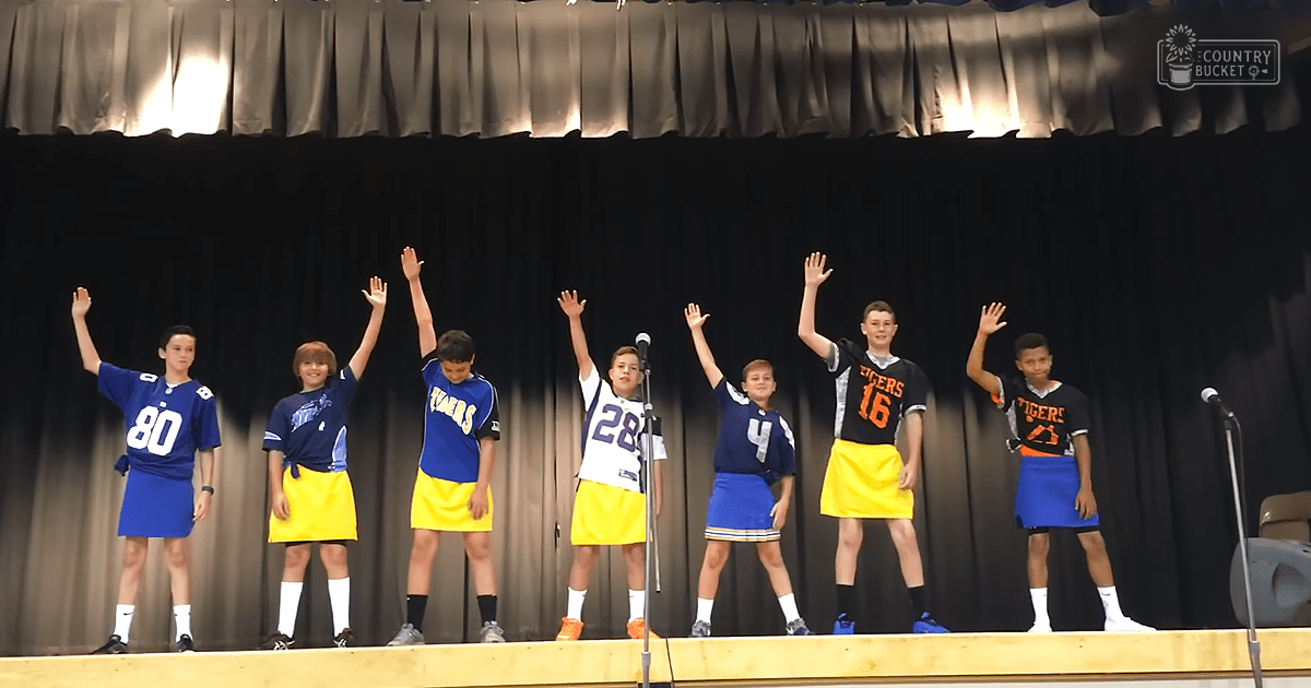 A group of children performing in a school auditorium or gymnasium, wearing blue and yellow jerseys with numbers and the word 'Tigers', on stage under artificial lighting.