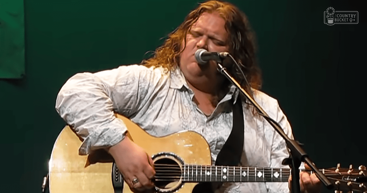 Adult male musician in relaxed long-sleeved button-up shirt, holding acoustic guitar on stage with dark green backdrop and 'Madly Odd' logo.