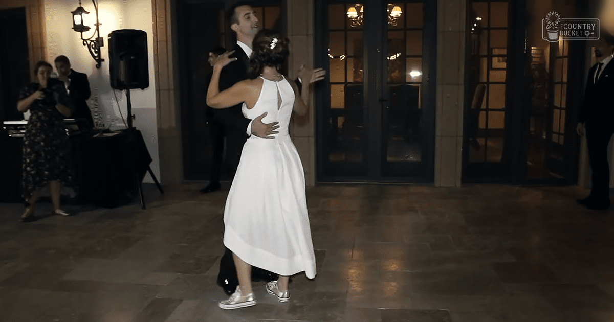A couple dancing indoors at a wedding reception with stone-tiled floors and neutral walls illuminated by artificial evening lighting.