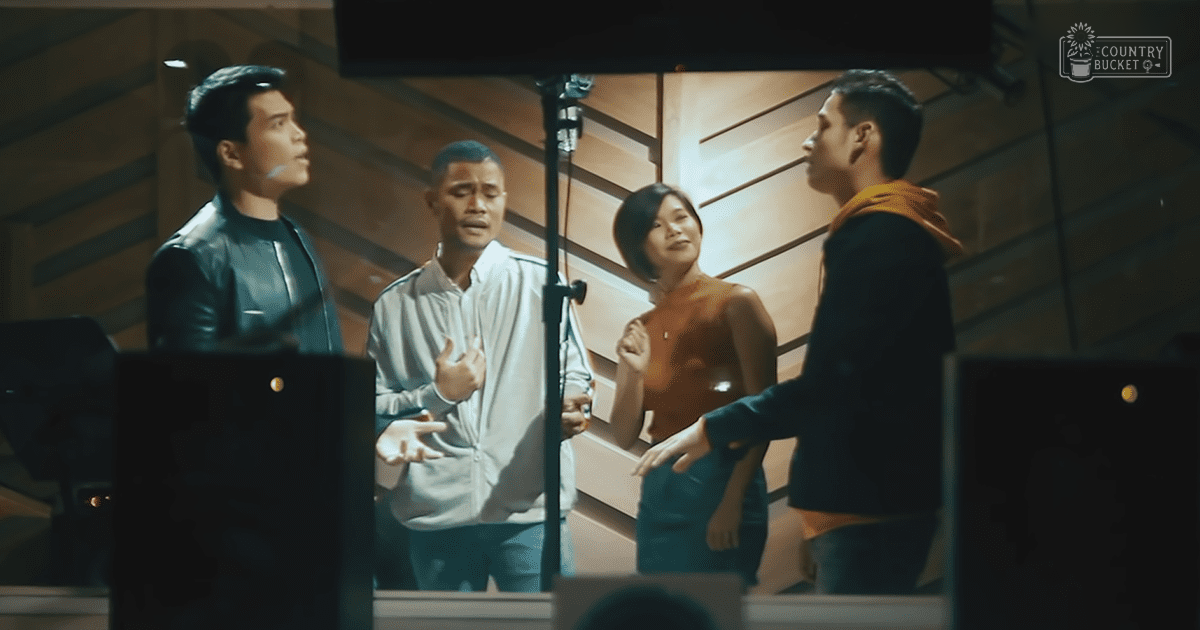 Four adults in an indoor studio or recording room with wooden paneling and audio equipment, wearing casual and semi-formal attire under artificial lighting.