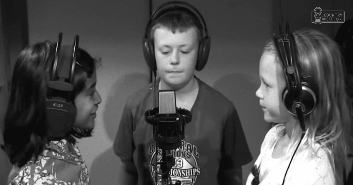A child wearing a floral-patterned dress stands with two other children, one in a dark T-shirt and another in a white top, all positioned near a microphone in what appears to be an indoor recording studio setting.