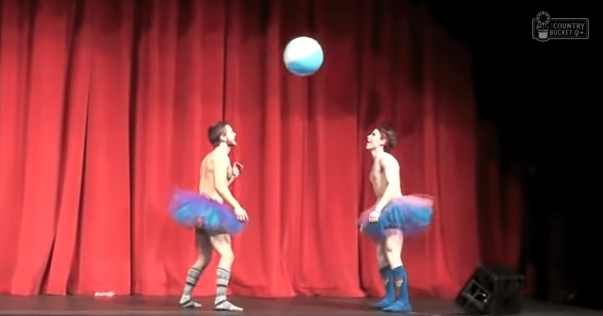A young adult male performer wearing blue tutus and knee-high ribbons-patterned socks stands on a stage with a red curtain backdrop in an indoor theater setting under focused lighting.