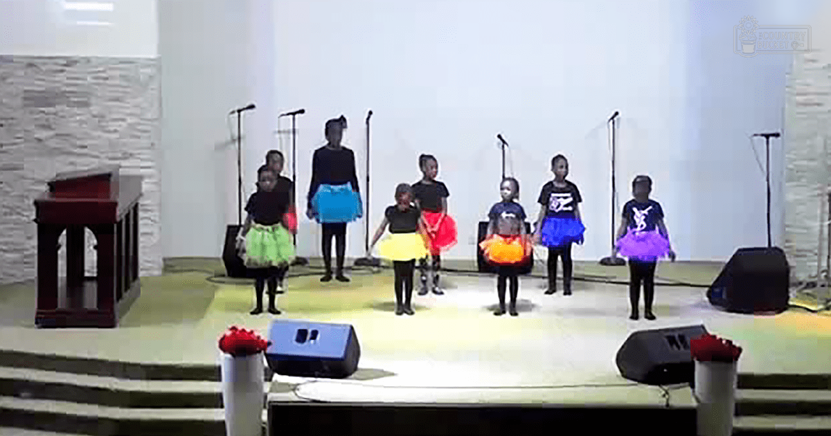 Children performing on stage at an indoor event with a wooden podium and microphones.
