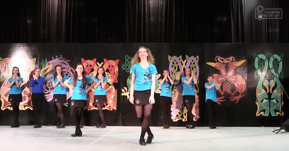 A young adult female dancer in a blue t-shirt and black skirt performing with a group of young adults dressed similarly at an indoor dance competition or performance.