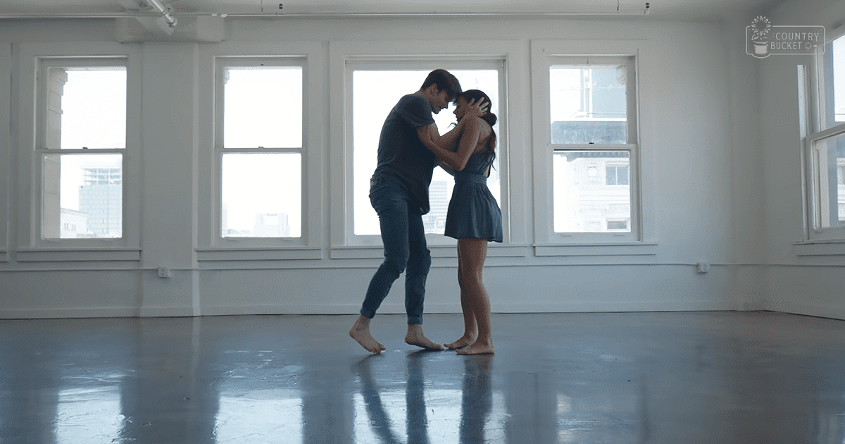 A young adult couple dancing in an empty room with large windows, bathed in natural light, wearing casual attire and a sheer dress.