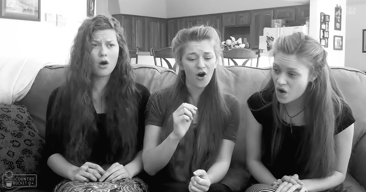 Three teenage girls wearing casual tops, sitting indoors with a living room and kitchen visible behind them.