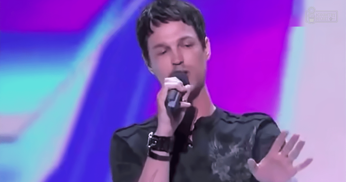 A young adult male singer performs on stage under vibrant purple, blue, and pink lights.