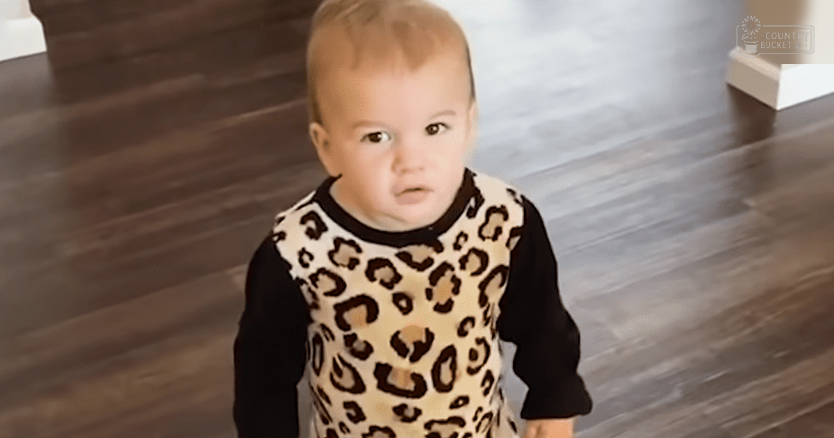 A toddler wearing a long-sleeved leopard print top with black sleeves stands indoors in daylight, surrounded by simple home decor.