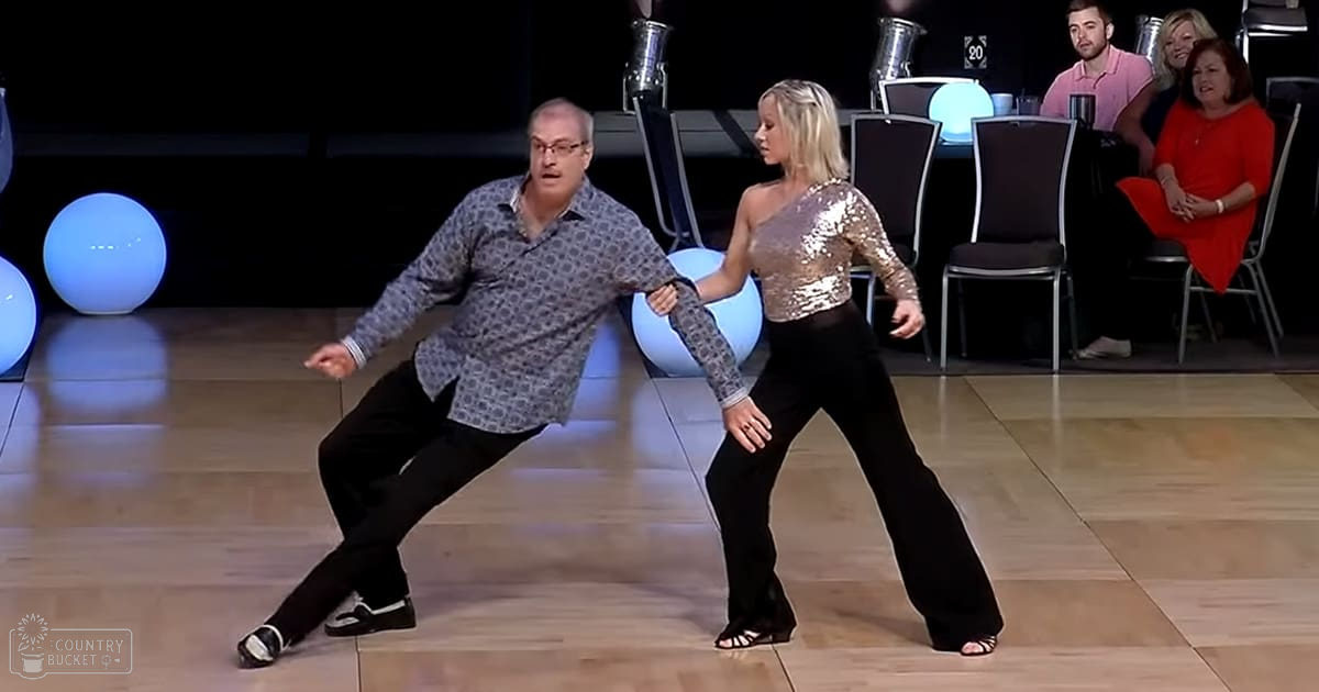 A dance performance scene in a studio setting with male and female dancers, audience members, round blue illuminated spheres on the floor, black chairs for the audience, and a dark backdrop with indistinct objects.