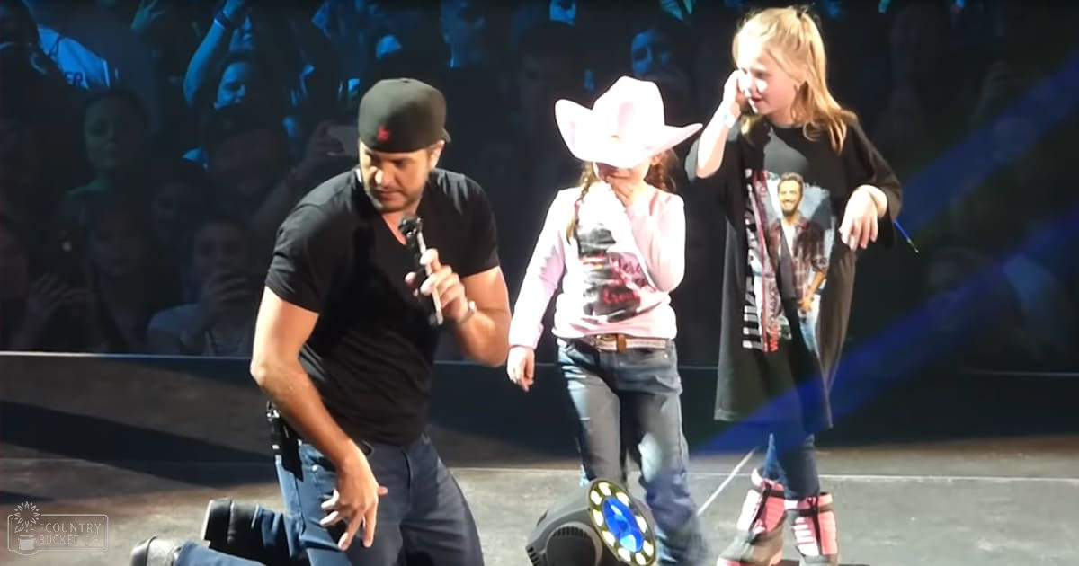 A young adult male singer performs on stage with two children, likely at an indoor concert venue. The performer wears a black T-shirt, dark jeans, and a backward cap; the children are dressed in colorful clothing and accessories.