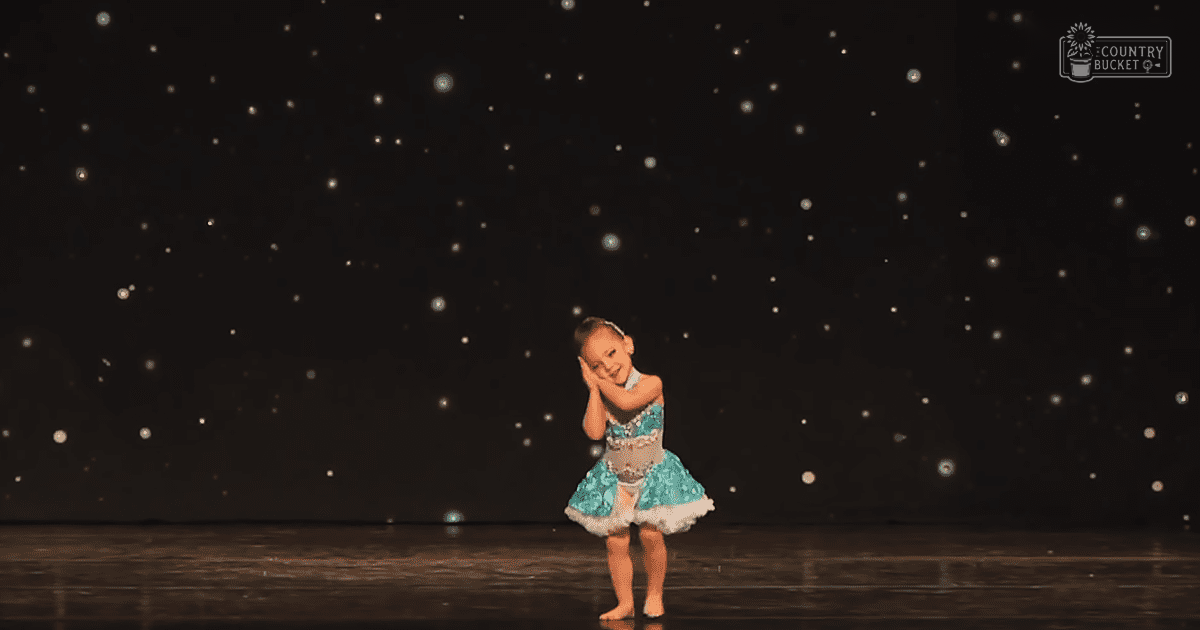 A toddler girl in a modern children's dance attire, barefoot on an indoor stage with a dark starry backdrop.