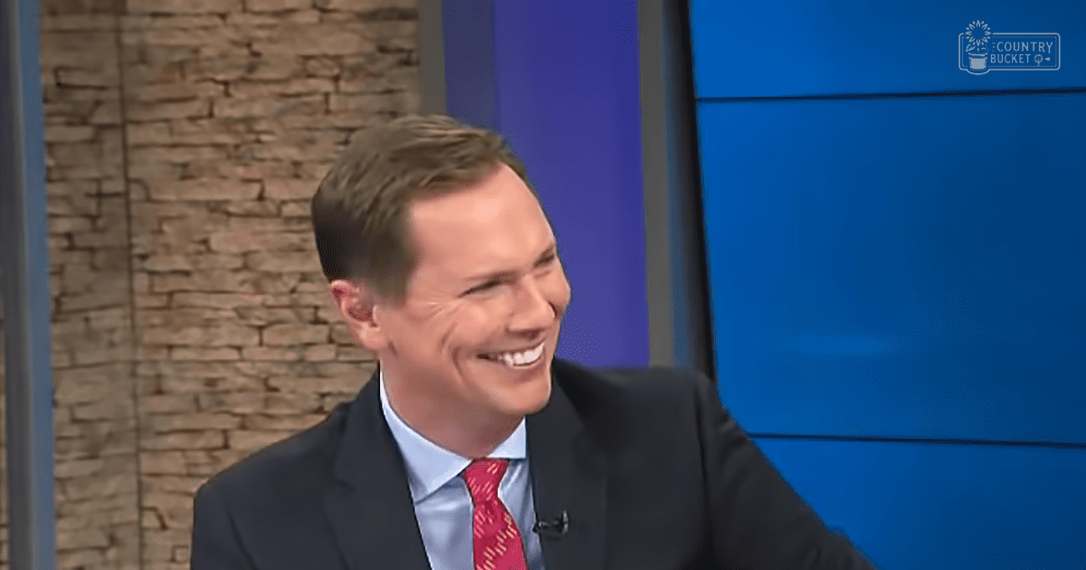 A man, adult male, in a formal dark suit with a light blue dress shirt and red patterned tie, standing indoors in what appears to be a television studio setting. He is wearing a small microphone clipped to his lapel.