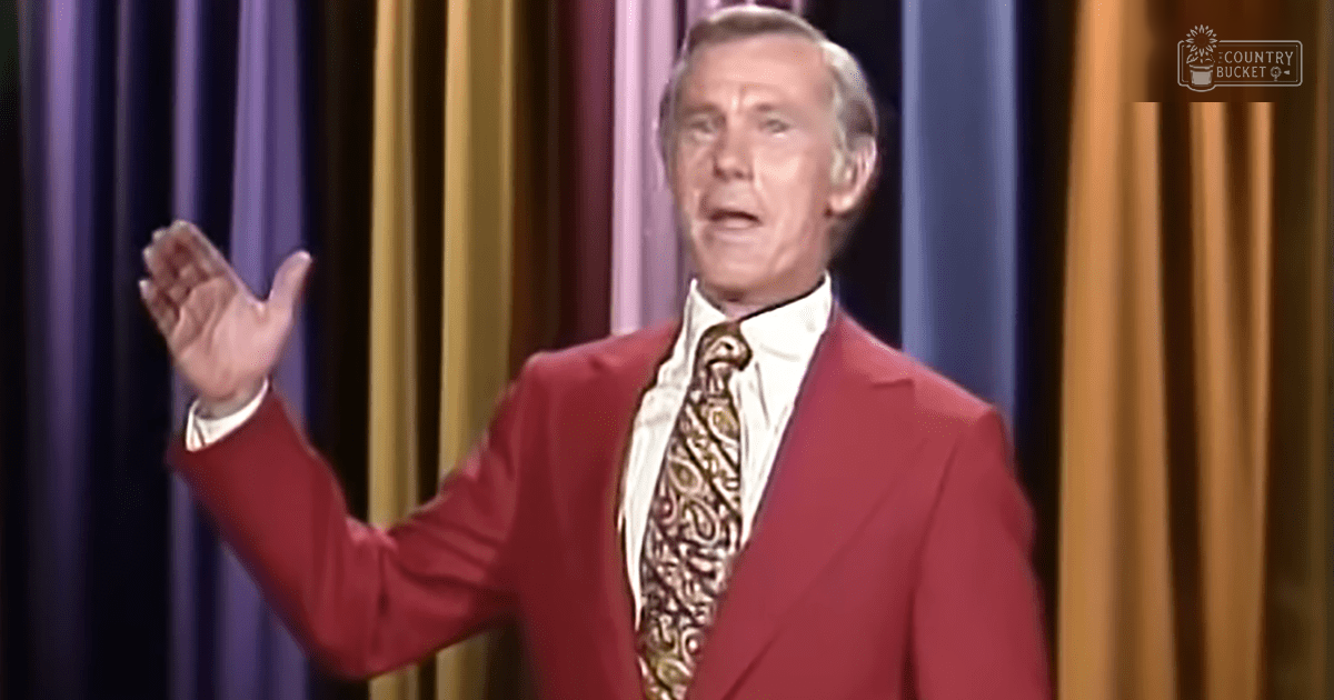 A middle-aged male performer in a red suit with a white shirt and patterned tie, performing on stage with purple and gold curtains.