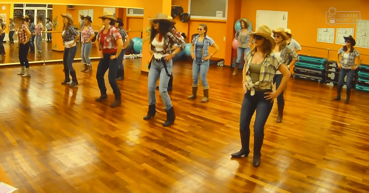 Young adult female dancers in a dance studio with wooden floors, mirrors on back wall reflecting them, yoga mats stacked to the right, and posters on walls. Orange walls, blue jeans, plaid shirts, white tops, brown boots.