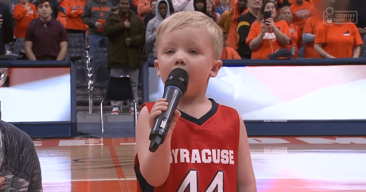 A young child wearing a red 'Syracuse' basketball jersey, holding a microphone at an indoor sports arena where adults in orange 'Syracuse' shirts are present.