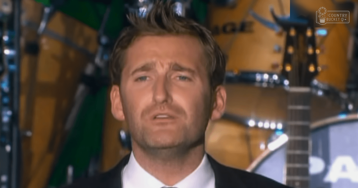 A man, possibly middle-aged with a dark suit and white dress shirt, stands indoors at what appears to be a music event. Musical instruments like guitars are visible behind him.