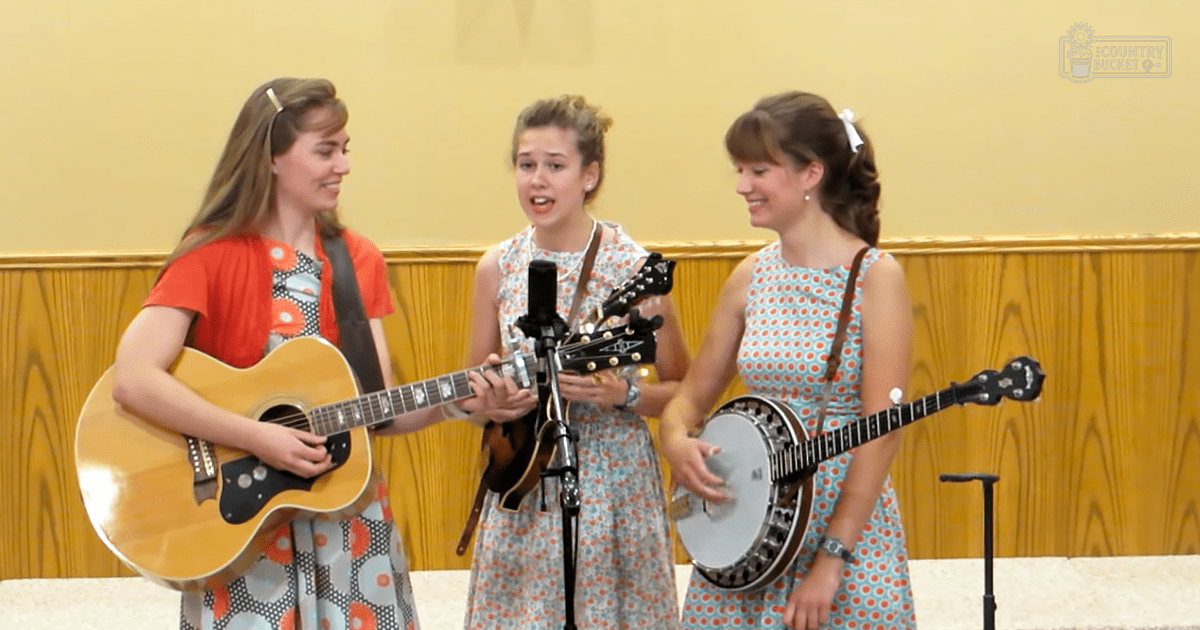 Young adult females performing a musical act indoors, wearing patterned dresses with floral and red dot patterns, surrounded by acoustic instruments and wooden panel walls.