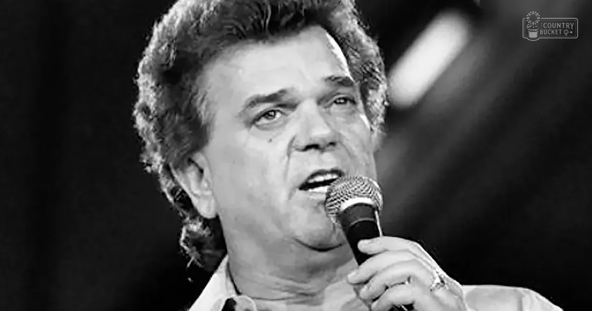 A middle-aged adult male singer wearing a white, relaxed-fit shirt with long sleeves and collar, performing into a microphone at an indoor venue under stage lighting.