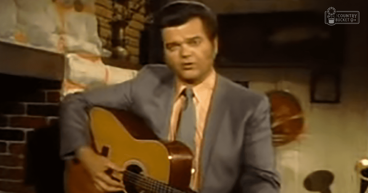 A man playing a guitar indoors, wearing a dark gray suit jacket and light pink dress shirt with matching tie, surrounded by warm indoor lighting and a brick fireplace.