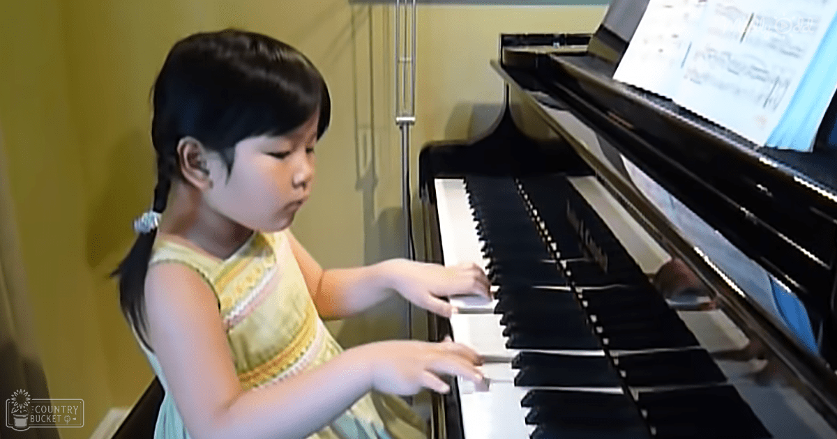 A young child wearing a yellow and orange patterned dress plays piano in an indoor setting with bright yellow walls.
