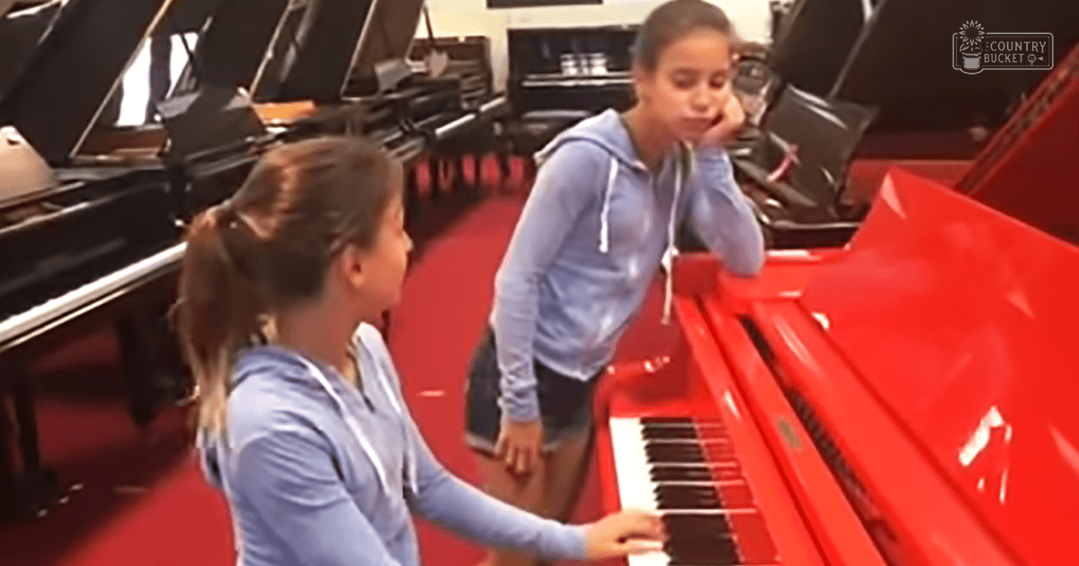 A young adult female and a child/teenage male play piano together in what appears to be a music store or studio, focusing on their light blue hoodies against the red carpeting with black grand pianos in the background.
