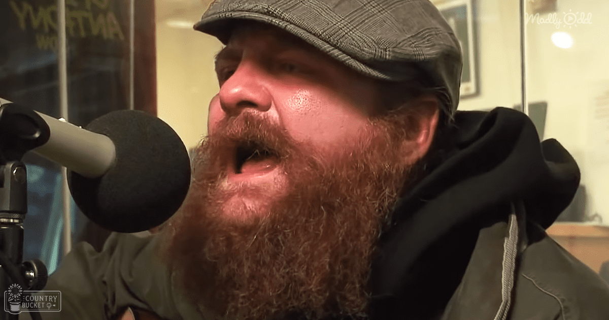A middle-aged adult male wearing a dark green jacket with a hood and a checkered flat cap stands indoors near a microphone in what appears to be a radio or podcast studio, surrounded by framed artwork and glass panels.