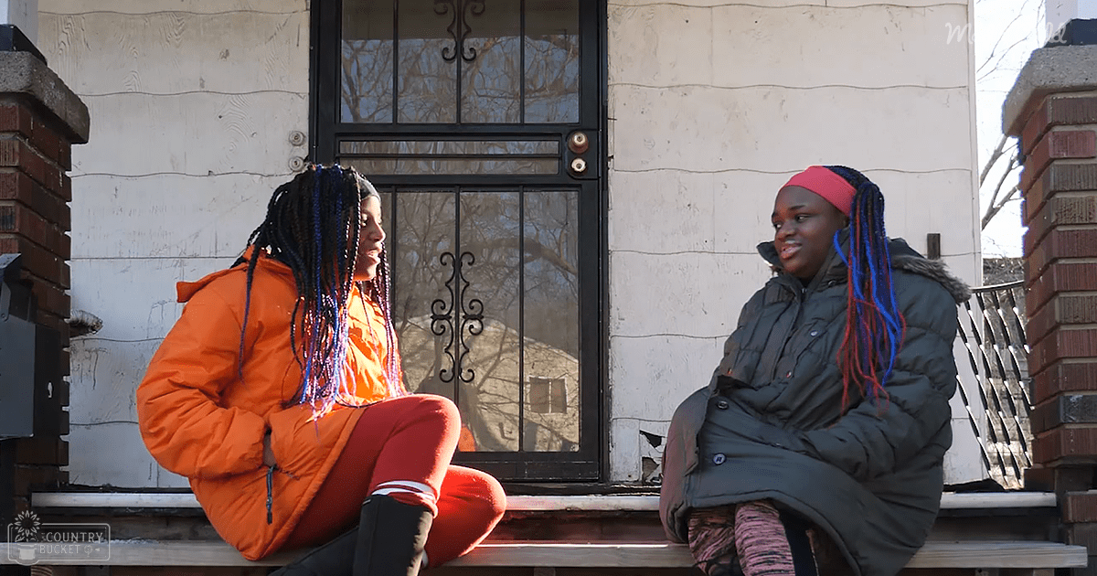 Two young adult females sitting outside a residential house, wearing bright orange and dark green puffer jackets in winter with natural daylight illuminating the scene.