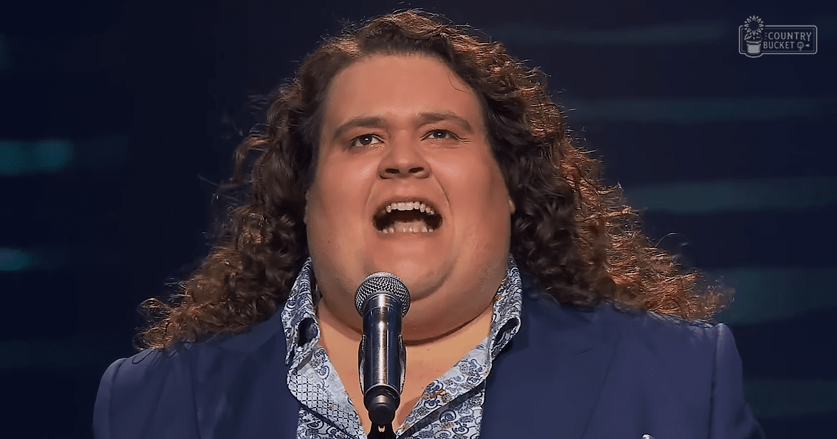 A young adult male in a modern casual or semi-formal attire with a blue button-up shirt with white and blue patterned design, singing into a microphone at what seems to be an indoor performance setting.