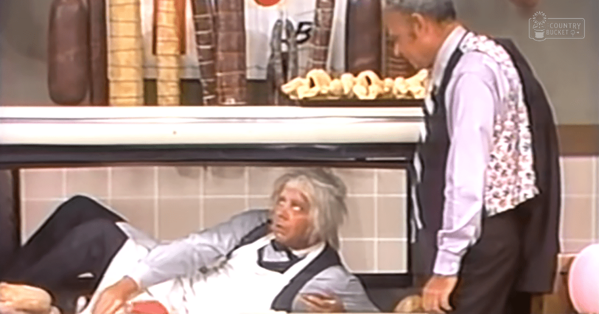 An older adult male in a diner setting, wearing a white shirt with a patterned dark vest over it. A standing adult male in a floral vest and formal pants is also present.